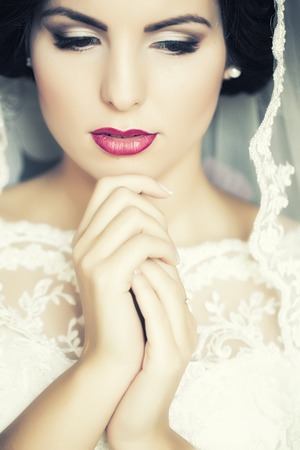 Portrait with side view of one beautiful young brunette sensual pensive bride in white lace dress and veil on head holding hands near face, vertical pictureの写真素材