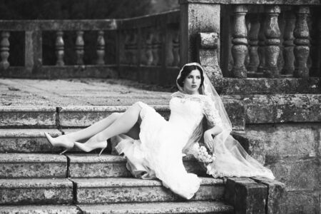 Full length view of one beautiful sensual pensive brunette female bride in long white lace wedding dress and veil and shoes sitting on stony stairs with of roses black and white, horizontal pictureの写真素材