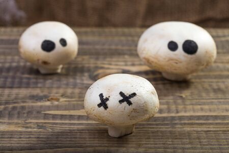 Photo still life set of three Halloween white button mushrooms champignons with ghost face eyes drawn in black felt pen standing on wooden table over blurred rustic background, horizontal pictureの写真素材