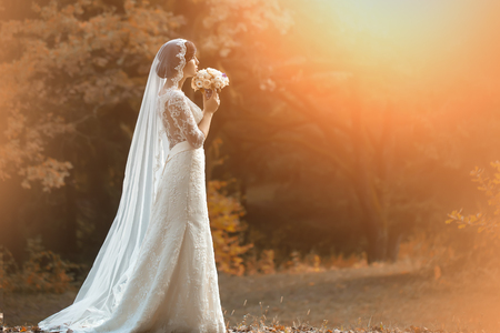 Full length side view of one beautiful sensual young brunette bride in long white wedding dress and veil standing in forest holding bouquet outdoor on natural background, horizontal pictureの写真素材