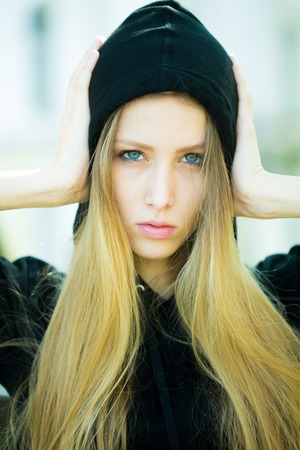 Portrait closeup of pretty blond girl with straight long hair flowed over her breast wearing black hood pin hands to head posing on streetscape background, vertical pictureの写真素材