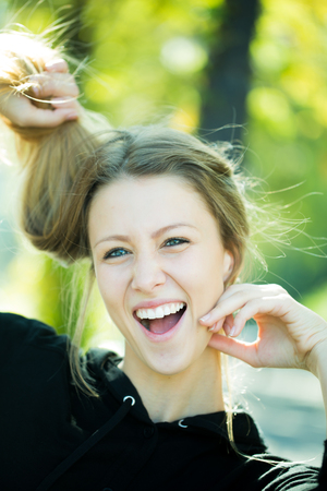 Portrait closeup of laughing pretty blond girl with long hair and big joyful smile taking hair in hands posing outside on blurred streetscape background, vertical pictureの写真素材