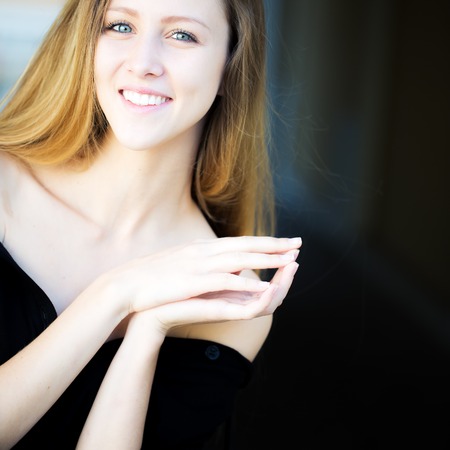 Portrait closeup of smiling pretty blond girl with long hair big joyful smile locking finger pads posing in black bare shoulders outside on blurred background, square pictureの写真素材