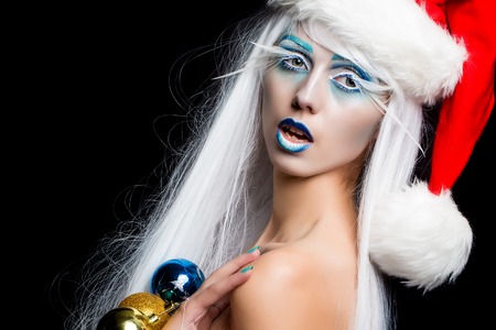One beautiful young fairy new year naked girl with winter blue makeup long white eyelashes and hair wig in red dress and christmas santa hat with decoration balls on black studio backdrop, horizontalの写真素材