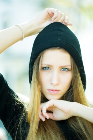 Portrait closeup of pretty blond girl with straight long hair flowed over her breast wearing black hood hands around face posing on streetscape background, vertical pictureの写真素材