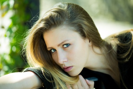 Portrait closeup of pretty blond girl with straight long hair looking at camera and posing  with head on stretched hand on blurred streetscape background, horizontal pictureの写真素材