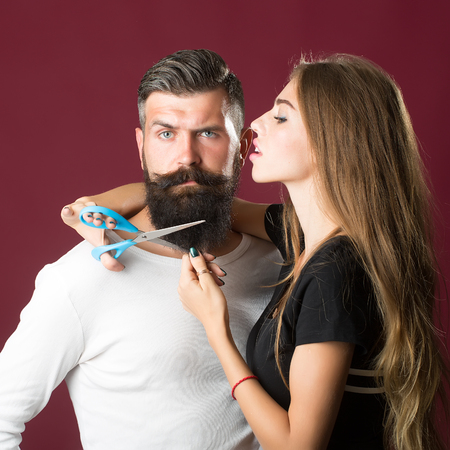 Closeup view of young beautiful couple of beautiful female barber cutting with scissors long beard of handsome man in studio on purple background, square pictureの写真素材