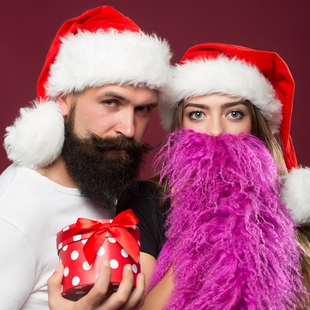 Young beautiful happy new year couple of man with long beard with present box and woman in red santa christmas hat with fur in studio on purple background, square pictureの写真素材