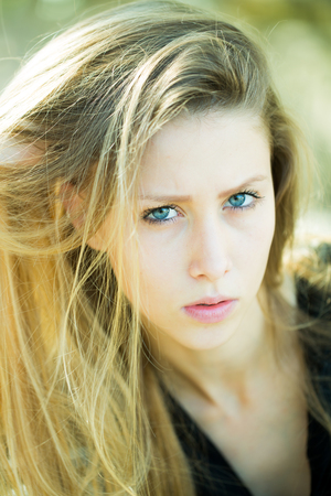 Portrait closeup of serious pretty blond girl with blue eyes long straight hair sidepart dishevelled looking towards camera posing outside on ligh background, vertical pictureの写真素材