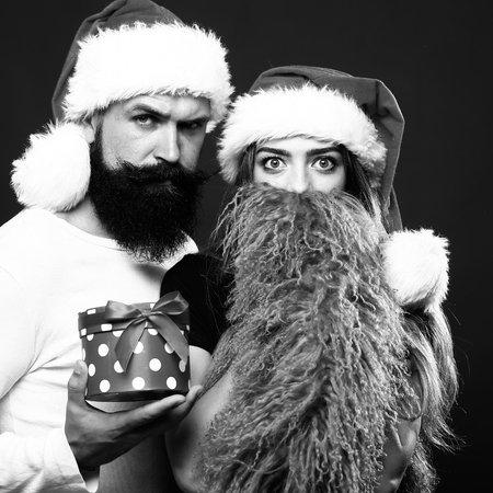 Young beautiful happy new year couple of man with long beard with present box and woman in red santa christmas hat with fur in studio black and white, square pictureの写真素材