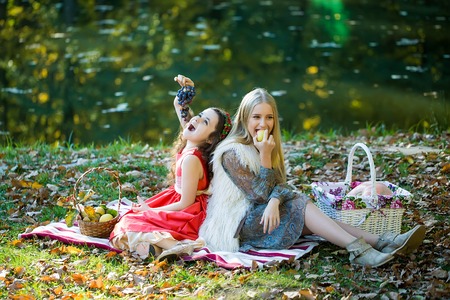 Two smiling girlfriends in beautiful clothes eating grapes and apple on plaid with fruits and vegetables picnic wicker baskets on lawn near lake in autumn park, horizontal photoの写真素材