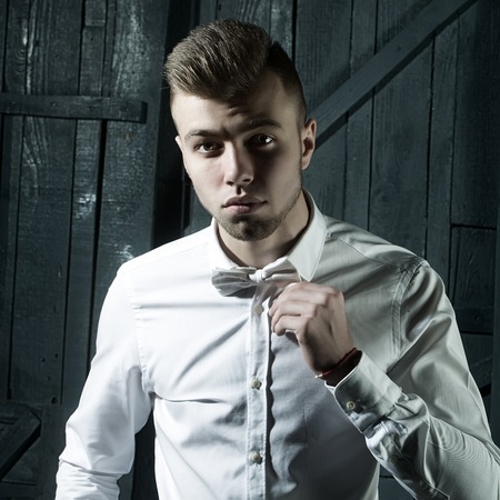 One sensual young handsome man with beautiful hairstyle in white shirt with bow standing in studio on wooden background, square pictureの写真素材