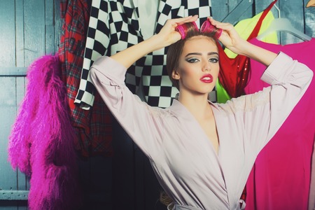 Closeup view of one beautiful young sensual housewife woman in dressing gown with hair-rollers on head standing in wardrobe among many bright clothes with raised hands, horizontal pictureの写真素材
