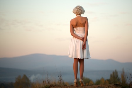 Full length back view of one young beautiful fashionable pensive retro sexy woman with blonde hair in white dress standing outdoor in dusk on sky natural background copy space, horizontal pictureの写真素材