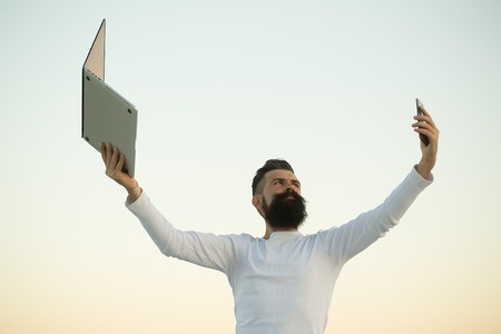 One handsome young bearded business man in white sweater holding and working on laptop with mobile phone outdoor on light sky natural background, horizontal pictureの写真素材