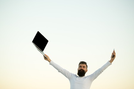 One handsome young bearded business man in white sweater holding and working on laptop with mobile phone outdoor on light sky natural background, horizontal pictureの写真素材