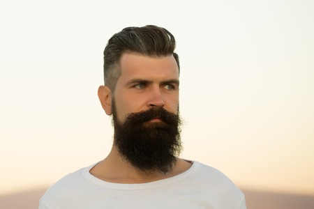 Closeup portrait of one handsome young stylish thoughtful man with long dark lush beard in white sweater looking away standing outdoor on light sky background, horizontal pictureの写真素材