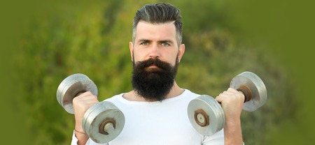 Closeup portrait of one handsome strong young man with long dark haired beard training with dumbells outdoor on green natural background, horizontal pictureの写真素材