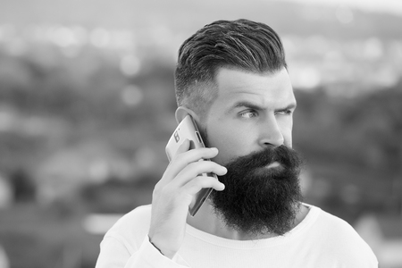Closeup portrait view of one handsome young pensive man with long dark haired beard speaking on mobile phone outdoor on blurred natural background black and white, horizontal pictureの写真素材