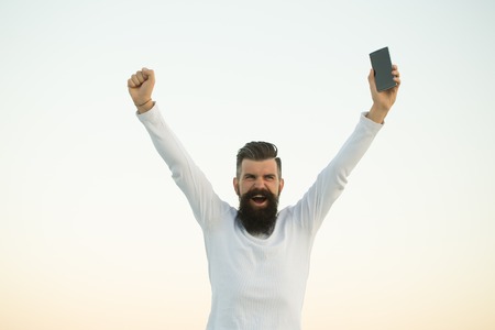 One handsome young bearded emotional business man in white sweater holding cell phone outdoor on light sky natural background, horizontal pictureの写真素材