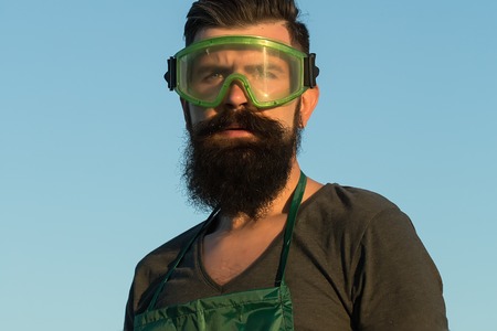 Closeup view of one handsome young adult man worker with long lush beard in green apron and work glasses standing outdoor in sunset on blue sky background, horizontal pictureの写真素材