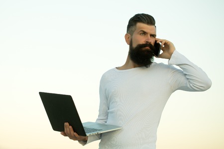 One handsome young bearded business man in white sweater holding and working on laptop talking on mobile phone outdoor on light sky natural background, horizontal pictureの写真素材