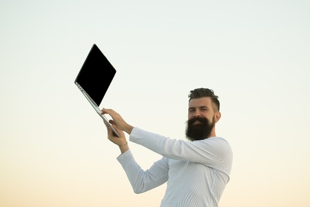 One handsome young bearded business man in white sweater holding and working on laptop outdoor on light sky natural background, horizontal pictureの写真素材