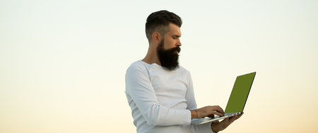 One handsome young bearded business man in white sweater holding and working on laptop outdoor on light sky natural background, horizontal pictureの写真素材