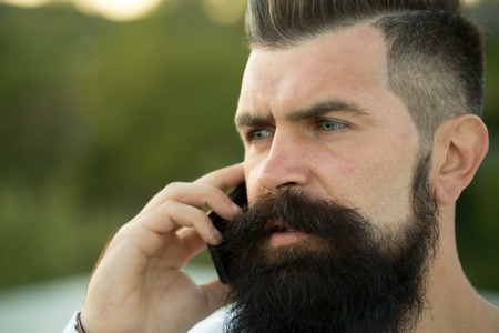 Closeup portrait view of one handsome young serious man with long dark haired beard speaking on mobile phone outdoor on blurred green natural background, horizontal pictureの写真素材