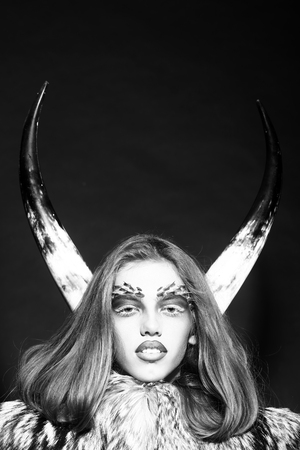 Closeup portrait of one beautiful wild young woman with bright animal monkey makeup with thorns on face and antlers in fur coat on black background black and white, vertical pictureの写真素材