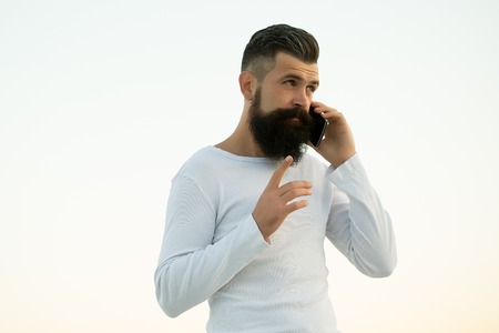 One handsome young bearded serious business man in white sweater holding and speaking on cell phone outdoor on light sky natural background, horizontal pictureの写真素材