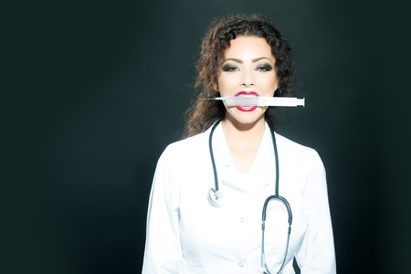 Closeup of one serious sexual beautiful brunette medical worker with stethoscope in white nurse uniform holding injecting suringe in mouth for botox shot or drugs in studio, horizontal pictureの写真素材