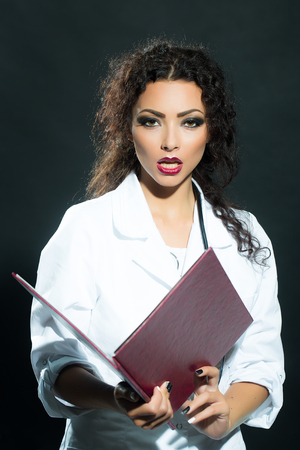 Closeup view portrait of one beautiful young sensual brunette sexy doctor with long curly hair stethoscope in white nurse hospital uniform holding big brown red medical card, vertical pictureの写真素材