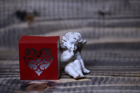Closeup view of one beautiful cupid angel decorative figurine near red paper greeting valentine box in shape of heart with no people on wooden background, horizontal pictureの写真素材