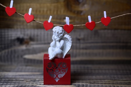 Closeup view of one beautiful cupid angel decorative figurine near red paper greeting valentine box near clothes-peg in shape of heart with no people on wooden background, horizontal pictureの写真素材