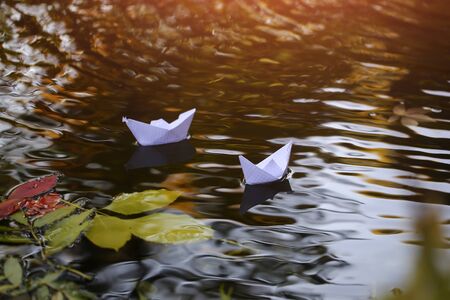 Closeup view of many beautiful colorful autumn tree leaves red yellow orange green colors floating on wavy water with reflection of nature with white paper ship on outdoor background, horizontal pictureの写真素材