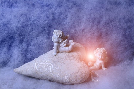 Closeup of beautiful soulful figurine composition of cupid angels for valentine day or christmas with small pillow in shape of heart lying on white wadding decorating snow with bright light sparkの写真素材