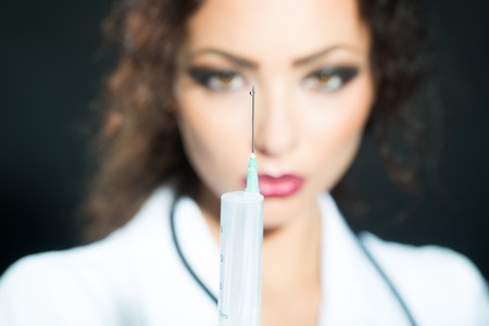 Closeup view blurred portrait of one serious sexual pretty medical worker in white nurse uniform holding injecting suringe for botox shot or drugs in studio, vertical pictureの写真素材