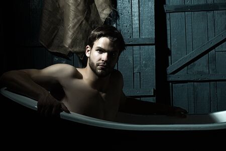 One handsome serious sensual sexy muscular young man with beautiful bare body sitting in white bath tub with foam taking shower looking forward indoor on wooden wall backdrop, horizontal pictureの写真素材