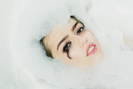 Closeup view portrait of one beautiful sensual sexy attractive young woman with wet bright makeup lying in bath tub with water and lots of white soap foam looking away, horizontal pictureの写真素材
