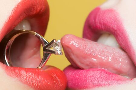 Closeup of female sexy lips with red lipgloss holding round beautiful gold wedding ring as new year present with gem in opne mouth with tongue and white teeth on yellow backdrop, horizontal photoの写真素材