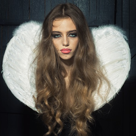 One beautiful tender dreaming young fashionable woman with long curly hair bright makeup and white fluffy angel wings with white feathers on back in black cloth dress on wooden background, squareの写真素材
