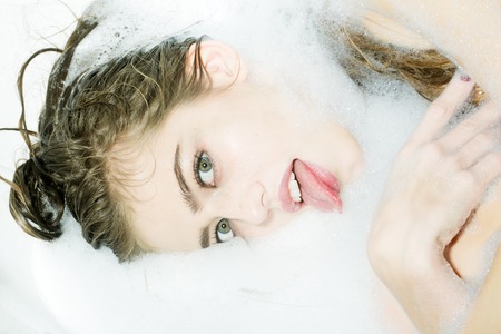 Closeup view portrait of one beautiful crazy flirtatious playful young woman with wet hair and bright makeup lying in bath tub with water and white soap foam looking away, horizontal pictureの写真素材