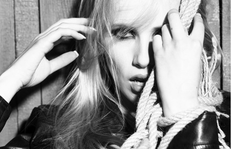 Portrait of one pretty sensual sexy young serious passionate blonde woman with long hair in leather black jacket with hands in rope on wooden background, horizontal pictureの写真素材