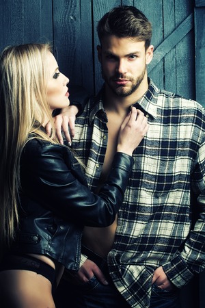 Young sexual couple of pretty blonde woman in leather jacket standing near handsome serious man in checkered shirt with bare muscular chest in studio on wooden background, vertical pictureの写真素材
