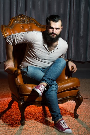 One stylish fashionable handsome brunette man with long lush beautiful beard in grey shirt and blue jeans sitting on brown leather retro chair looking forward indoor on studio background, vertical picの写真素材