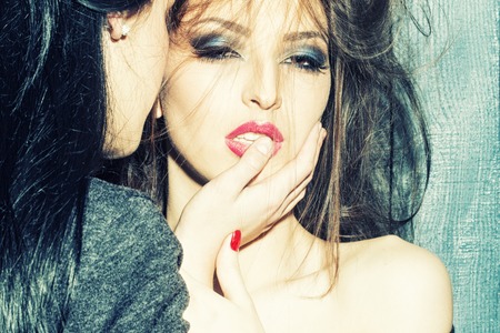 Closeup of two pretty sensual young brunette girls mistress with bare shoulder and hand in mouth embracing and kissing standing near wall indoor in studio, horizontal photoの写真素材