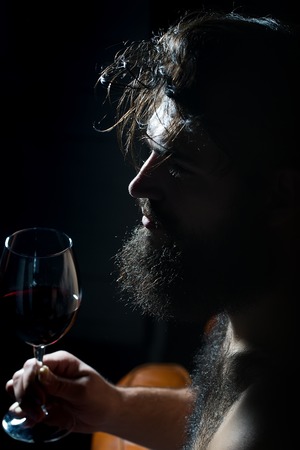 Closeup side view of one handsome sad pensive young adult man with long black lush beautiful beard and moustache holding glass with red wine indoor on blurred background, vertical pictureの写真素材
