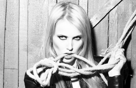 Portrait of one attractive sensual sexy young cool passionate blonde woman with long hair in leather black jacket holding rope in hand on wooden background, horizontal pictureの写真素材