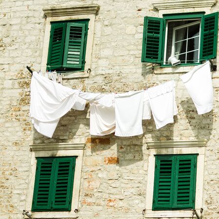 Photo closeup of white clean washed laundry linen on line to dry outside building air with green shutters bedding on sunny day on cityscape background, square pictureの写真素材